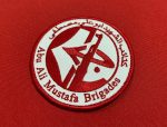 The image features a red and white embroidered patch with Arabic text and the name "Abu Ali Mustafa Brigades" written in English. The patch includes a graphic of a stylized weapon, symbolizing the group's identity. The background is a solid red fabric, highlighting the intricate details of the patch.