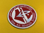 Red circular patch with white embroidery, featuring an abstract emblem combining a flag and a firearm, surrounded by Arabic script and the text "Abu Ali Mustafa Brigades." The patch represents the armed wing of the Popular Front for the Liberation of Palestine (PFLP).
