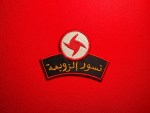 A red embroidered SSNP “Nusour al-Zawba’a” Eagles of the Whirlwind patch with the red-and-white whirlwind emblem above gold Arabic text lies on a bright red fabric background, photographed flat for a product listing on a collectible military insignia shop.