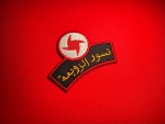 A red embroidered SSNP “Nusour al-Zawba’a” Eagles of the Whirlwind patch with the red-and-white whirlwind emblem above gold Arabic text lies on a bright red fabric background, photographed flat for a product listing on a collectible military insignia shop.