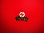 A red embroidered SSNP “Nusour al-Zawba’a” Eagles of the Whirlwind patch with the red-and-white whirlwind emblem above gold Arabic text lies on a bright red fabric background, photographed flat for a product listing on a collectible military insignia shop.