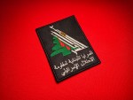 Lebanese Resistance Brigades patch featuring crossed rifles, dagger, green cedar tree, and Arabic text on a black background, displayed on a red fabric surface.