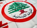 Lebanese Forces flag with green cedar tree emblem inside red circle, Arabic text “القوات اللبنانية,” and four red stylized crosses on white background, Christian militia political party Lebanon banner.