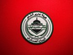 Circular embroidered patch with green satin border featuring Arabic text “هيئة تحرير الشام” and “كتيبة خالد بن الوليد – الجناح العسكري,” with a central shield showing a domed structure and rifle motif on a red fabric background