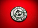 Circular embroidered patch with green satin border featuring Arabic text “هيئة تحرير الشام” and “كتيبة خالد بن الوليد – الجناح العسكري,” with a central shield showing a domed structure and rifle motif on a red fabric background