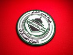 Circular embroidered patch with green satin border featuring Arabic text “هيئة تحرير الشام” and “كتيبة خالد بن الوليد – الجناح العسكري,” with a central shield showing a domed structure and rifle motif on a red fabric background