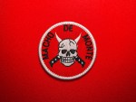 Embroidered circular Macho de Monte patch with a red background, white satin border, horned skull at center, crossed blades beneath the skull, and black “Macho de Monte” text around the edge.