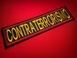 Embroidered CONTRATERRORISMO rectangular patch with gold lettering on black background and red satin border, photographed on red fabric background.