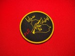Round black embroidered Saraya Awliya al-Dam patch with gold border, Arabic text, Iraq map outline, and raised fist holding a rifle on a red fabric background.