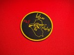 Round black embroidered Saraya Awliya al-Dam patch with gold border, Arabic text, Iraq map outline, and raised fist holding a rifle on a red fabric background.