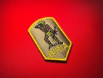 Angled view of the embroidered BIRIA Atlacatl reproduction patch on red fabric, showing the warrior design, yellow lettering, and satin edge.
